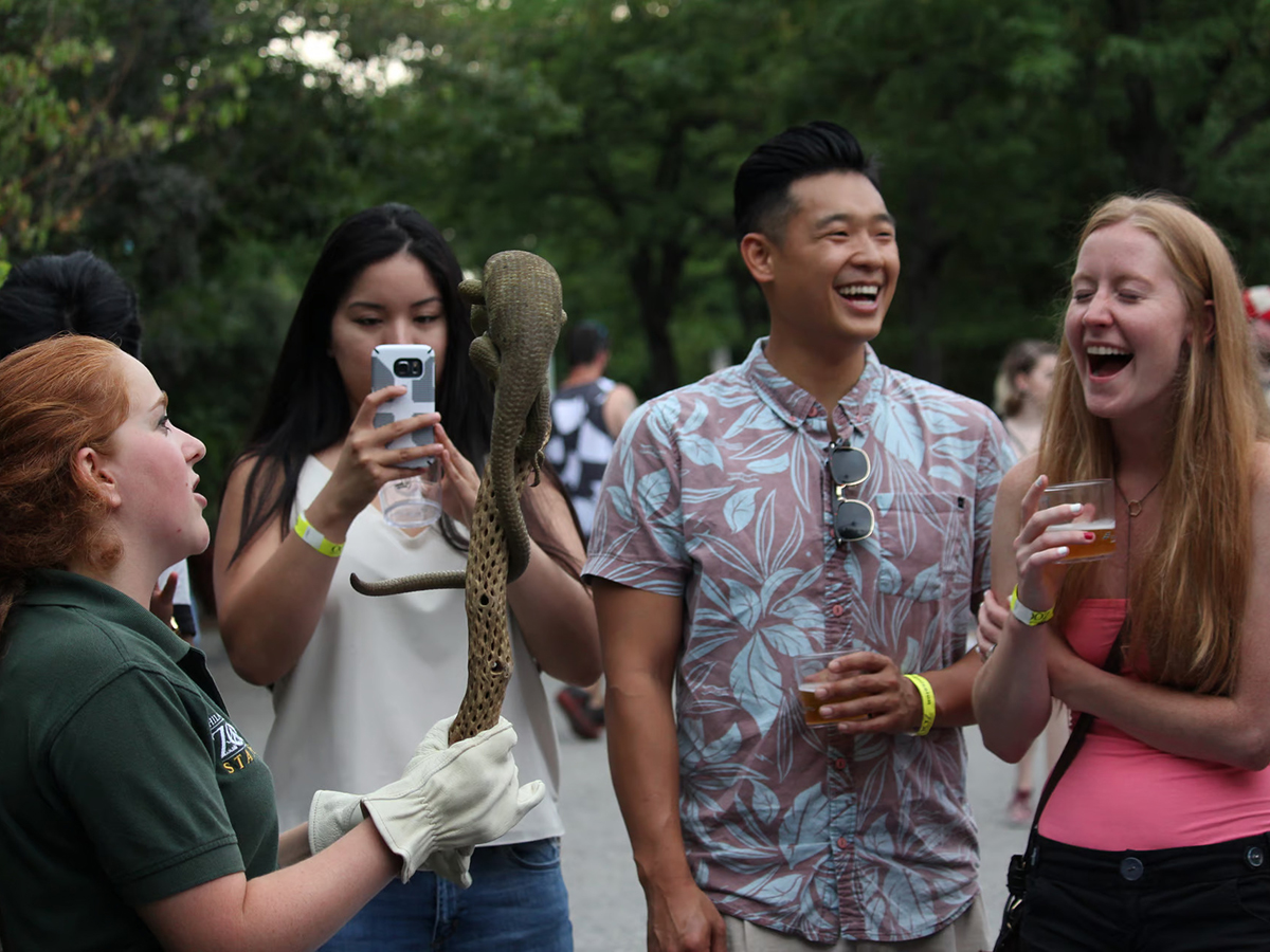 A group of smiling young adults holding cups of beer interact with a zookeeper holding a lizard at the Philadelphia Zoo's Ale Festival.
