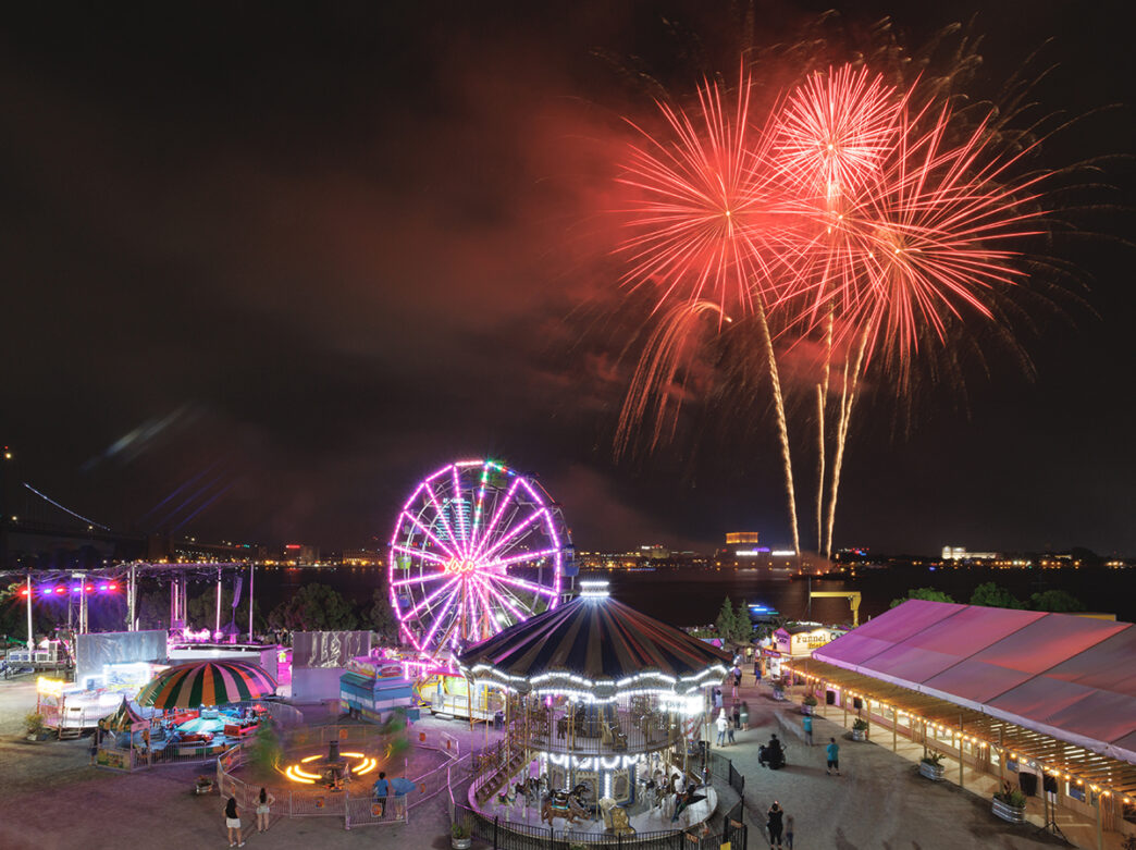 Vibrant red fireworks light up the sky over colorful carnival rides, including a glowing Ferris wheel at Independence Blue Cross RiverRink Summerfest.