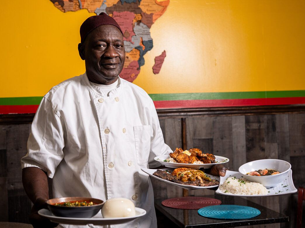 A chef in a white chef shirt smiles at the camera while holding plates of West African dishes at African Small Pot. Behind him is a yellow wall with a map of Africa on it.
