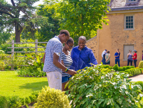 A group of people explore a lush garden on a sunny day at Bartram's Garden, with historic stone buildings in the background.