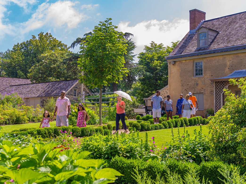Visitors stroll through the lush gardens at Bartram's Gardens, surrounded by greenery and historic buildings on a sunny day.