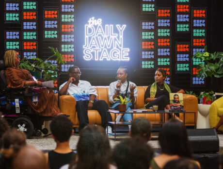 A group of panelists speak on stage at BlackStar Film Festival's Daily Jawn Stage in front of a live audience, framed by plants and vibrant neon sign.
