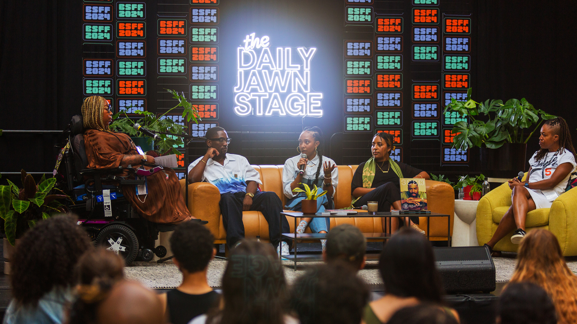 Un groupe de panélistes s'exprime sur la scène du Daily Jawn Stage du BlackStar Film Festival devant un public, encadré par des plantes et un néon vibrant.