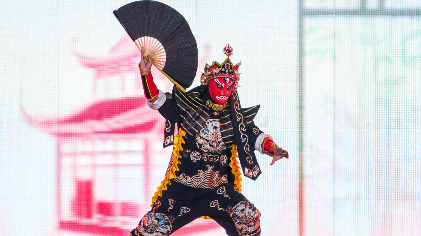 Um artista com uma roupa ornamentada em preto e dourado e uma máscara vermelha se apresenta no palco do Festival das Lanternas Chinesas.