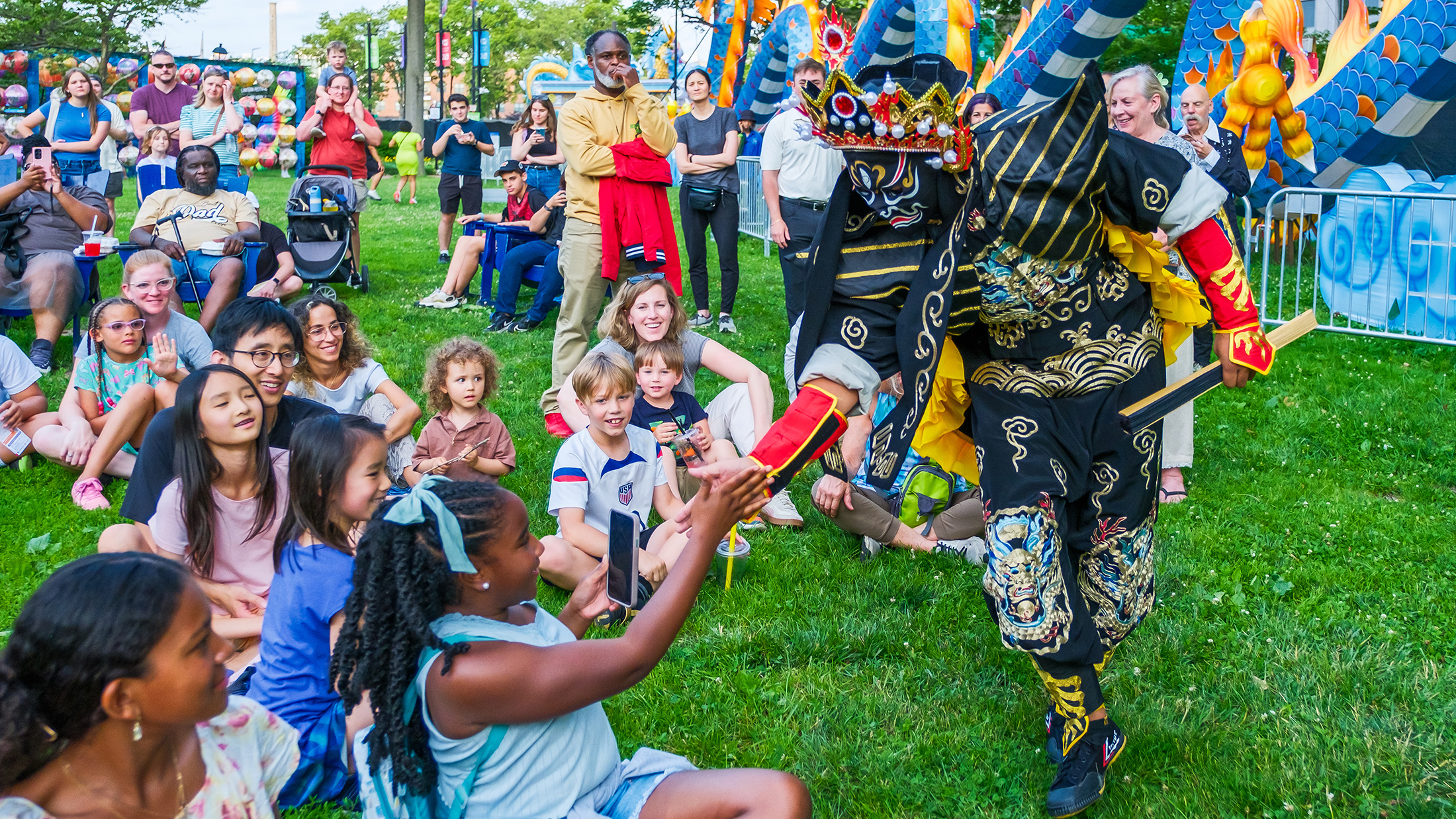 A costumed face-changer performer interacts with smiling kids seated on the grass during a lively show at the Chinese Lantern Festival.