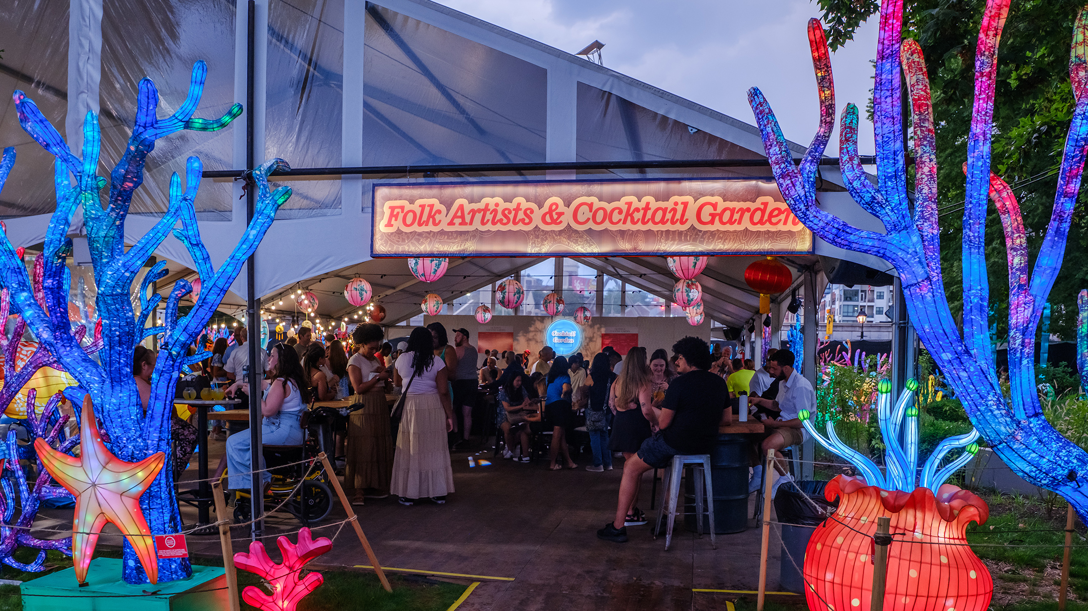 Visitors gather under a tent marked "Folk Artists & Cocktail Garden" surrounded by glowing coral and starfish lanterns.