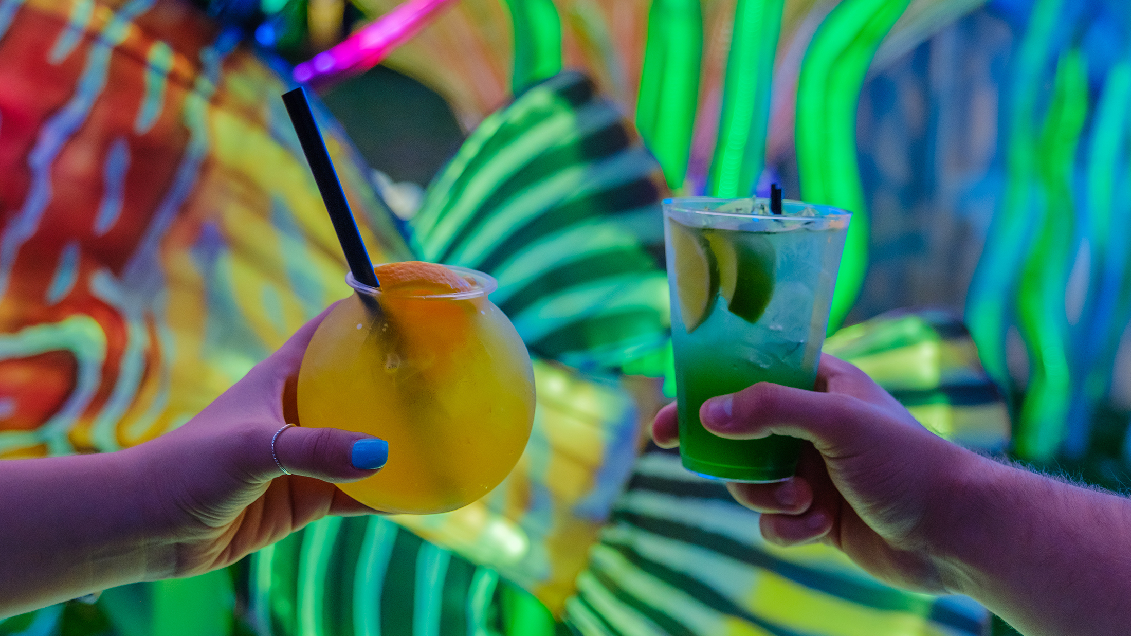Two colorful cocktails are held up by hands in front of a glowing lantern display.
