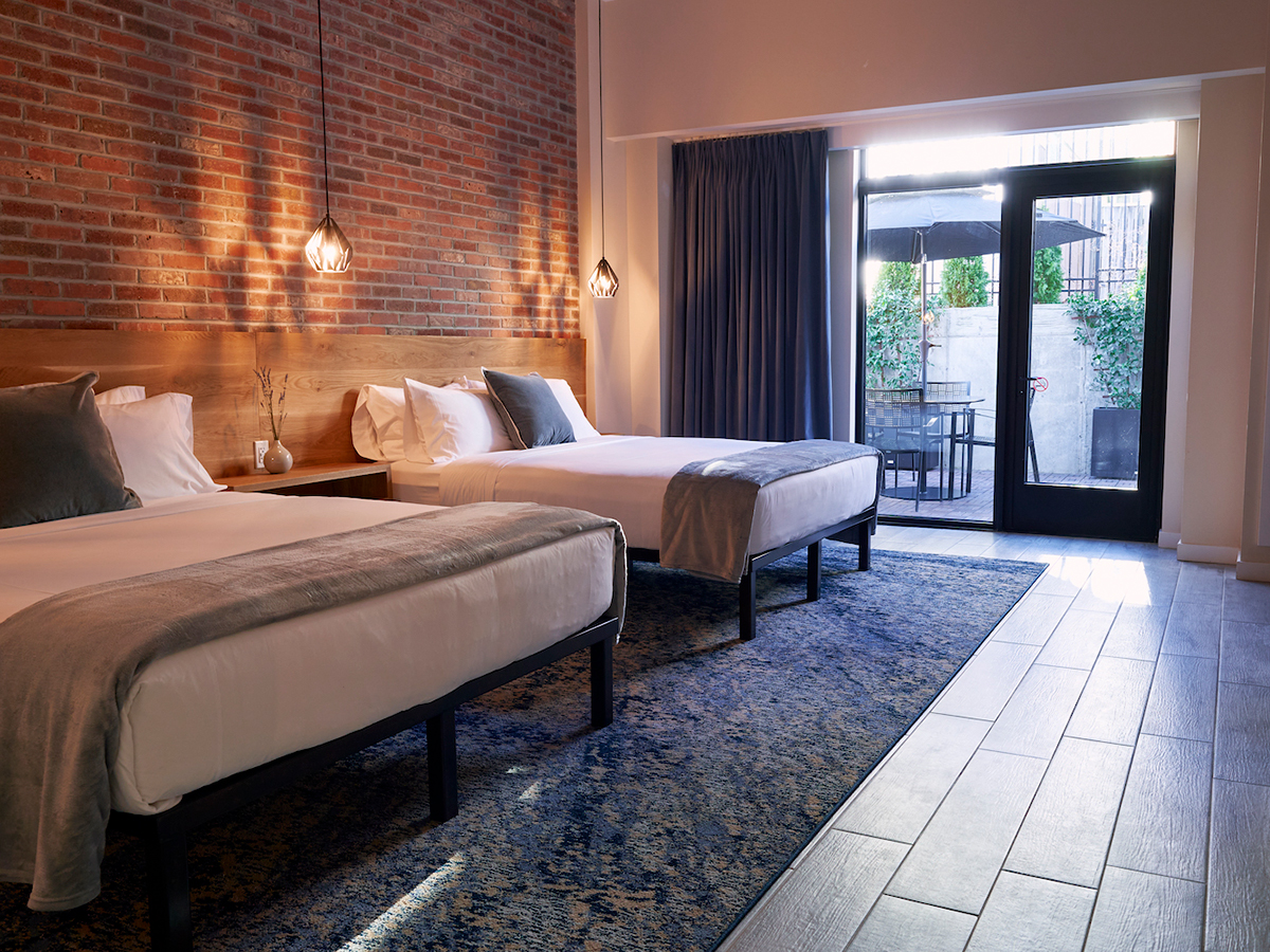 A guest room at featuring two queen beds with white linens against a brick wall at the Maj in Philadelphia. Pendant lights hang on either side of the bed and sliding glass door looks out to a patio.