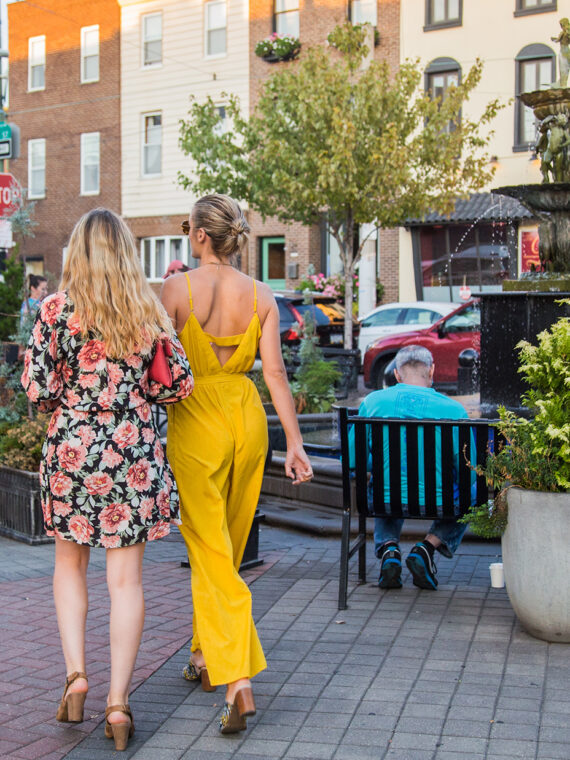 Two women walk past the Singing Fountain on East Passyunk Avenue, surrounded by shops, restaurants and people enjoying the neighborhood.