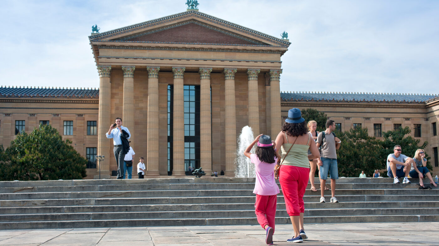 Visitors walk up the iconic stone steps outside the Philadelphia Museum of Art. The massive building, with tall columns and triangular pediment, rides in front of them as people sit and stand around the fountain the courtyard.