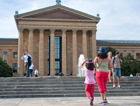 Visitors walk up the iconic stone steps outside the Philadelphia Museum of Art. The massive building, with tall columns and triangular pediment, rides in front of them as people sit and stand around the fountain the courtyard.