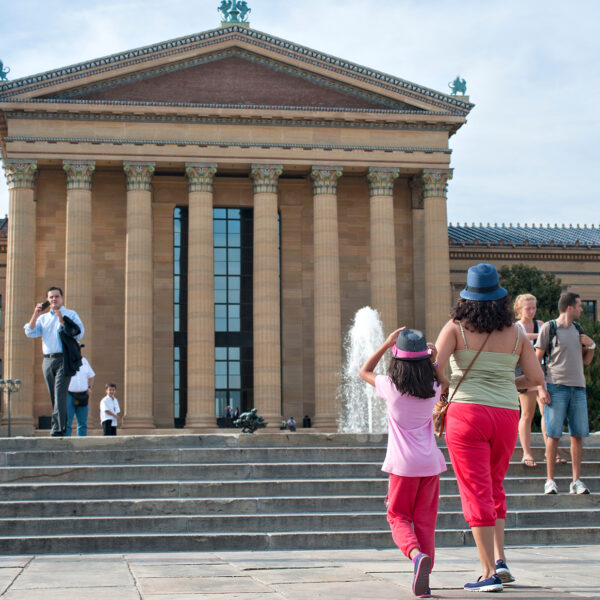 Visitors walk up the iconic stone steps outside the Philadelphia Museum of Art. The massive building, with tall columns and triangular pediment, rides in front of them as people sit and stand around the fountain the courtyard.
