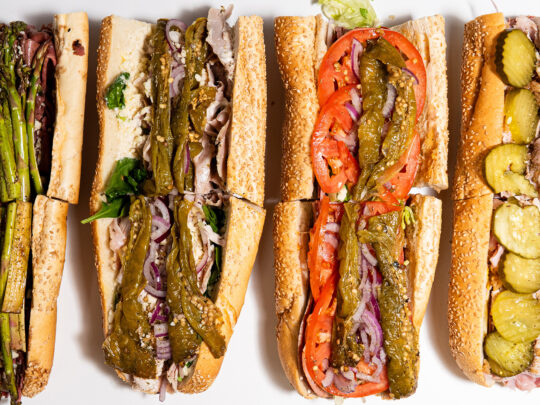 An overhead view of four hoagies from Fink's Hoagies, each filled with a variety of meats, cheeses, vegetables and toppings on sesame seed rolls.