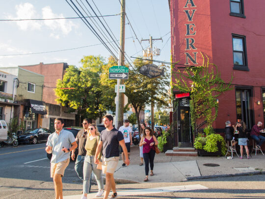 People cross a street in the heart of Fishtown, passing local bars and restaurants on a sunny day.