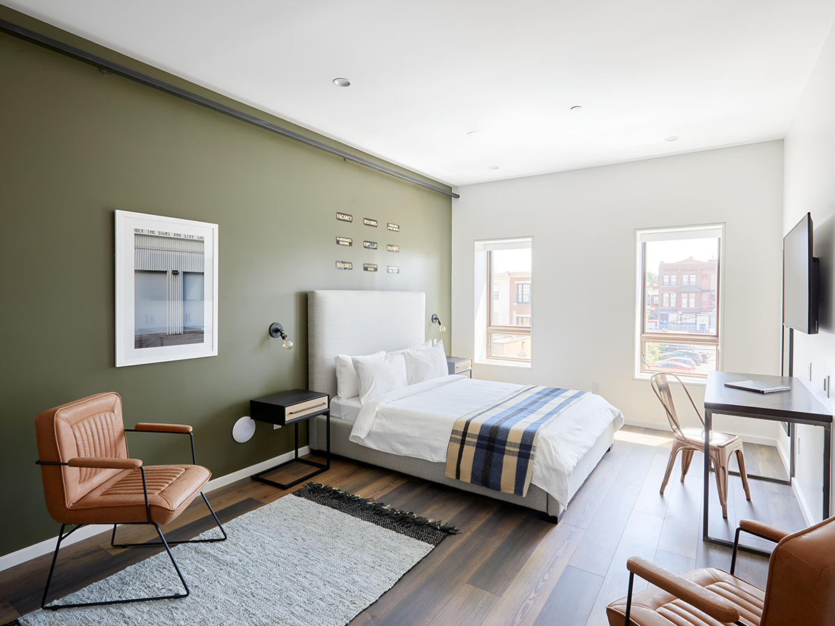 A modern guest room featuring minimalist decor, a bed with white linens and a striped blanket, leather chairs and large windows.