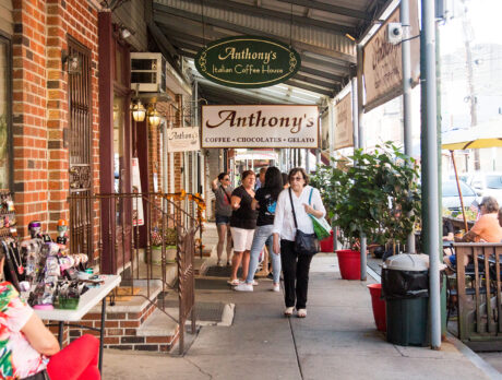People walk along the sidewalk at the Italian Market in Philadelphia. Signs for Anthony's Chocolate House hang from an overhang over the sidewalk.
