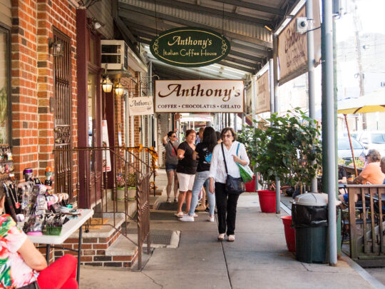 People walk along the sidewalk at the Italian Market in Philadelphia. Signs for Anthony's Chocolate House hang from an overhang over the sidewalk.