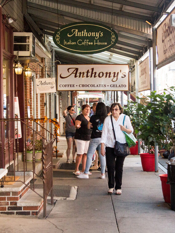 People walk along the sidewalk at the Italian Market in Philadelphia. Signs for Anthony's Chocolate House hang from an overhang over the sidewalk.