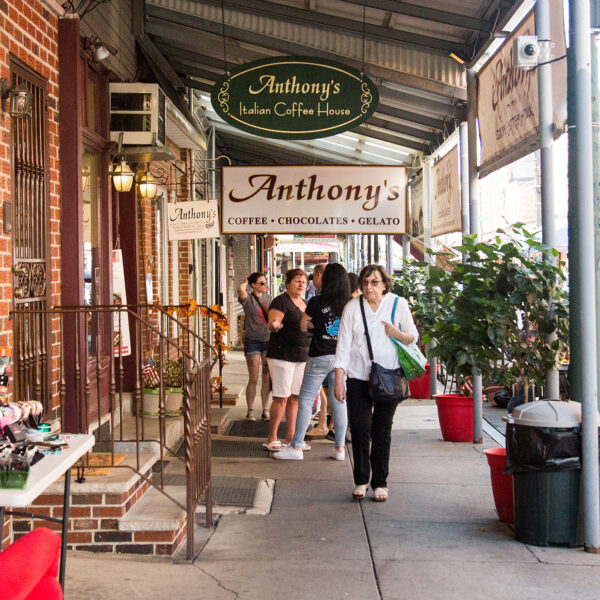 People walk along the sidewalk at the Italian Market in Philadelphia. Signs for Anthony's Chocolate House hang from an overhang over the sidewalk.