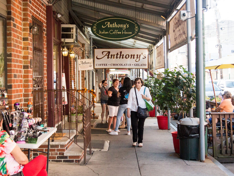 La gente camina por la acera en la Italian Market de Filadelfia. De un saliente sobre la acera cuelgan carteles de Anthony's Chocolate House.