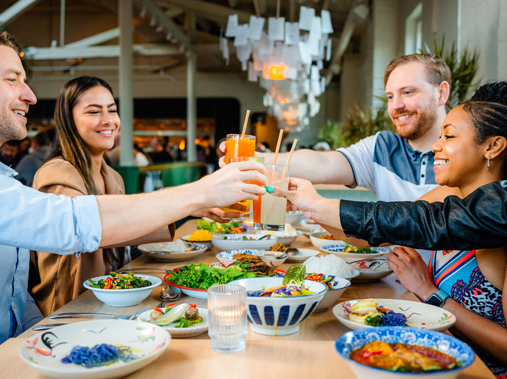 A group of smiling friends raise their glasses for a toast over a long table filled with colorful Thai cuisine.
