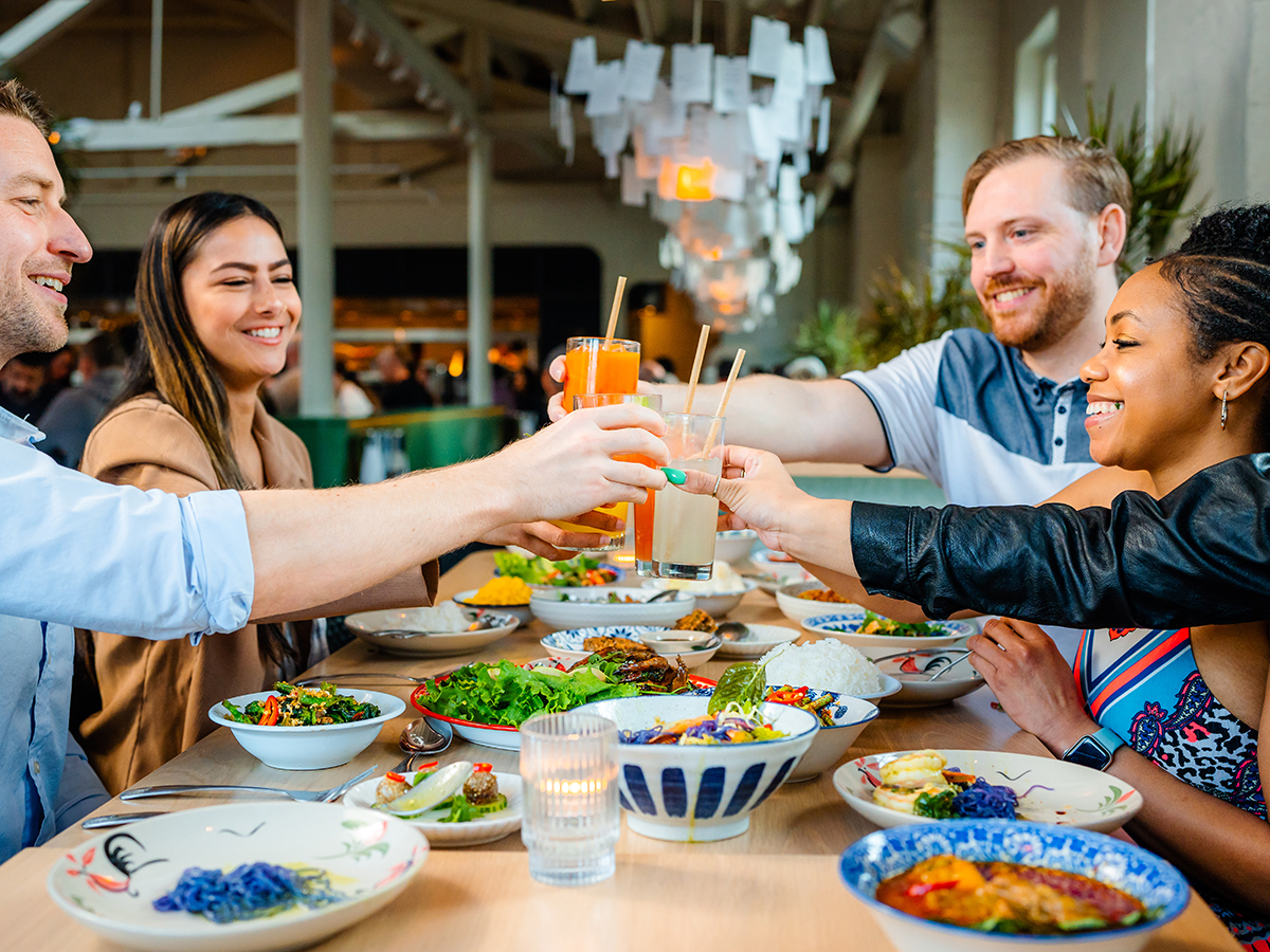 A group of smiling friends raise their glasses for a toast over a long table filled with colorful Thai cuisine.