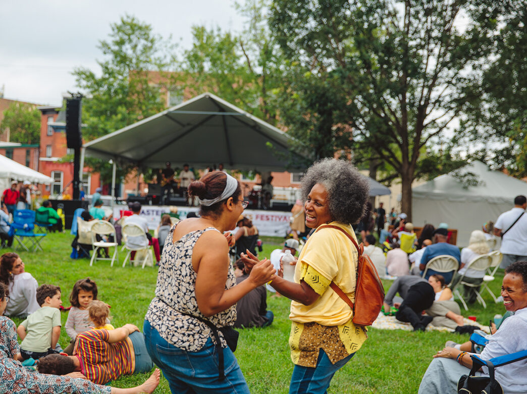 Twee vrouwen lachen en dansen in een park vol gras tijdens een levendig muziekfestival in de open lucht.