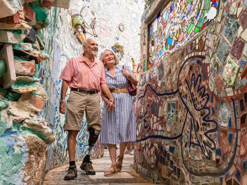 Um homem vestindo bermuda e camisa polo rosa segura a mão de uma mulher usando um vestido despojado e sandálias. Juntos, eles caminham pelas paredes de mosaico do Magic Gardens da Filadélfia.