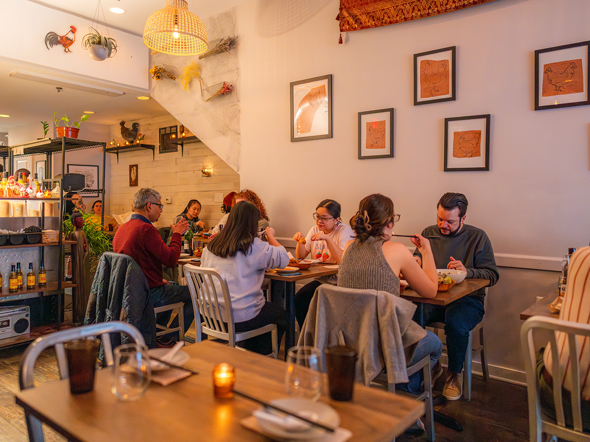 A warm, inviting dining room at Mawn with guests seated at small wooden tables, enjoying their meals. Drawings of chickens on brown paper are displayed in frames on the walls.