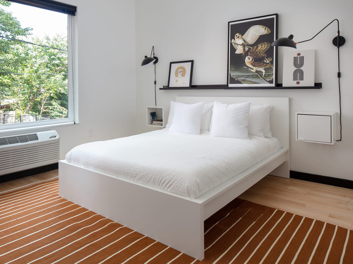 A modern hotel room with a queen bed made with white linens, a orange striped rug, paintings above the bed and a large window overlooking green trees.