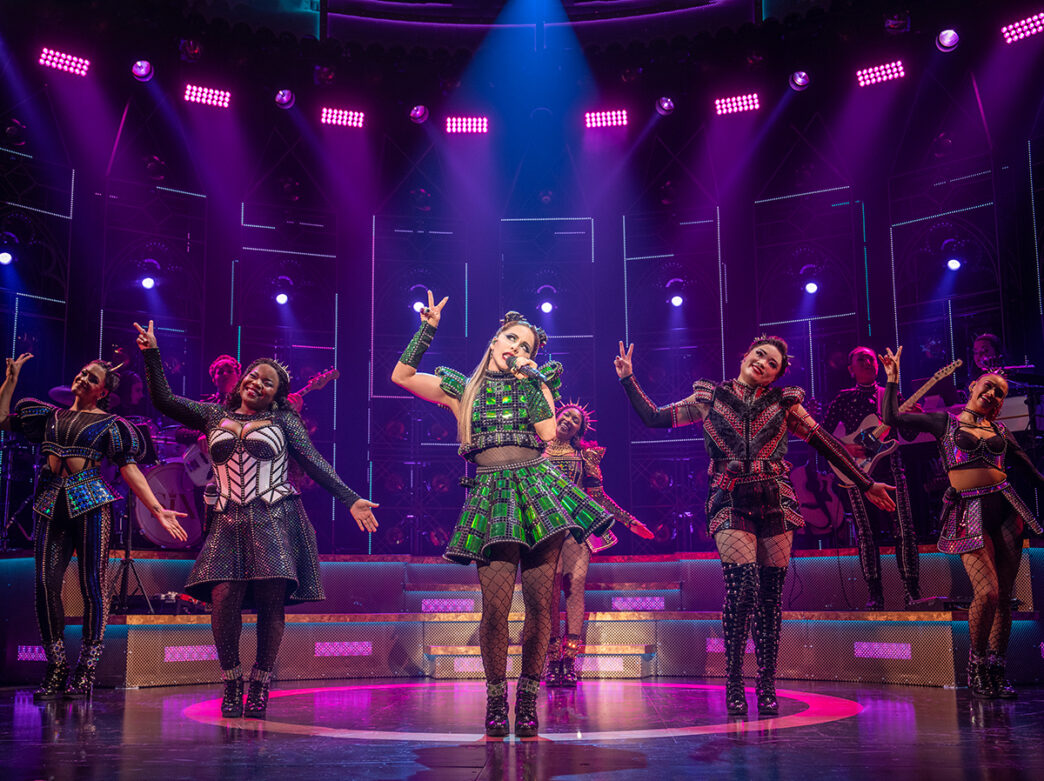 Cast members of SIX dance and sing on stage while wearing glittering costumes under dramatic stage lighting.