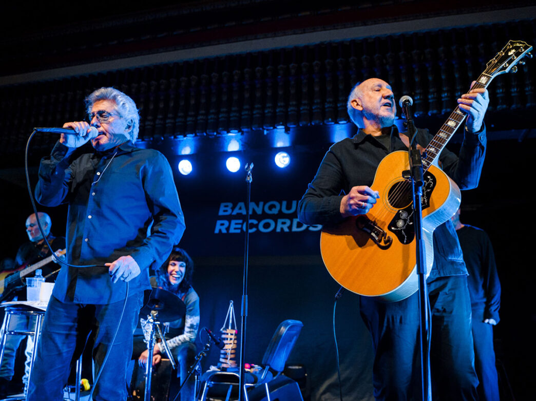 Two band members stand front and center on stage during a concert, one sings into a microphone and another plays the acoustic guiar.