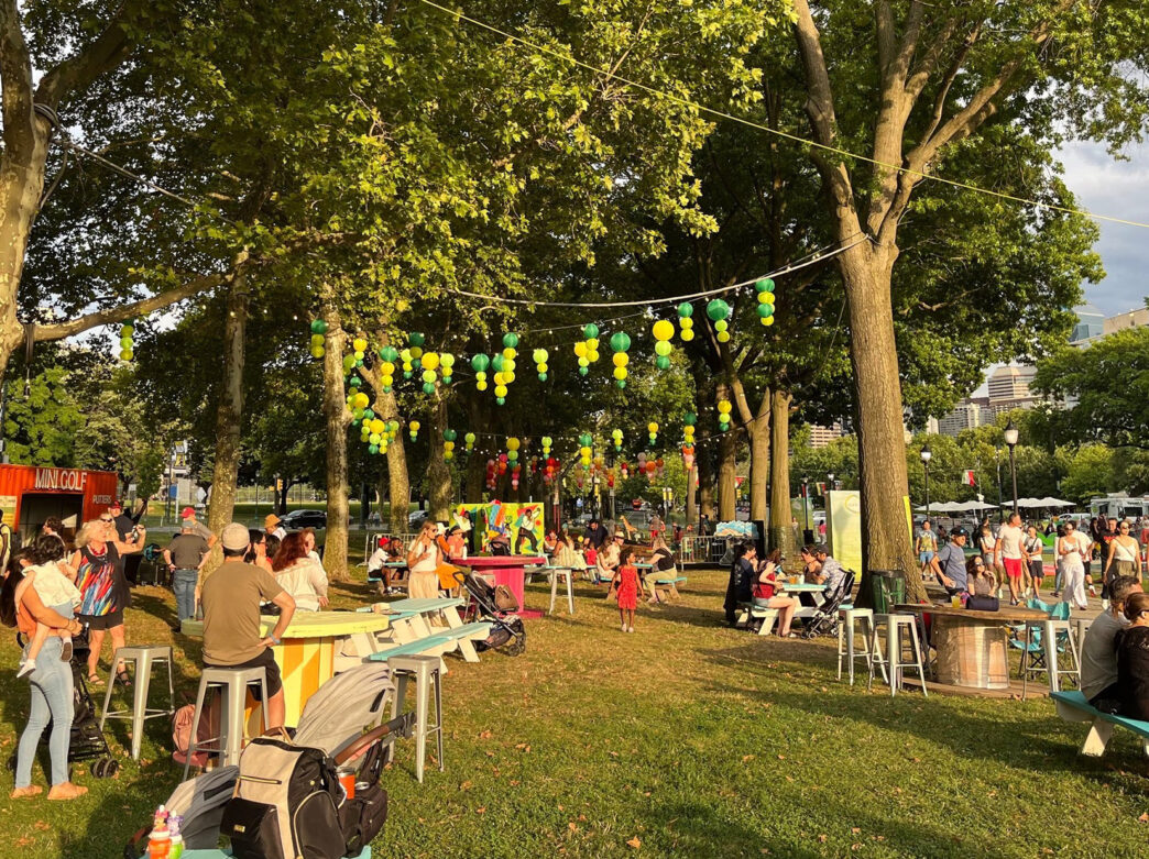 Groups of people relax at colorful picnic tables under green and yellow lanterns strung between trees at an outdoor beer garden.