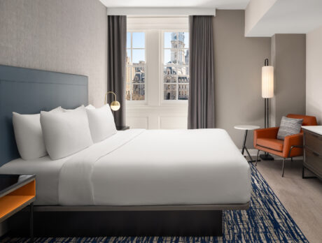 A hotel guestroom with a king bed with white linens, a orange accent chair and a window overlooking Philadelphia's City Hall.