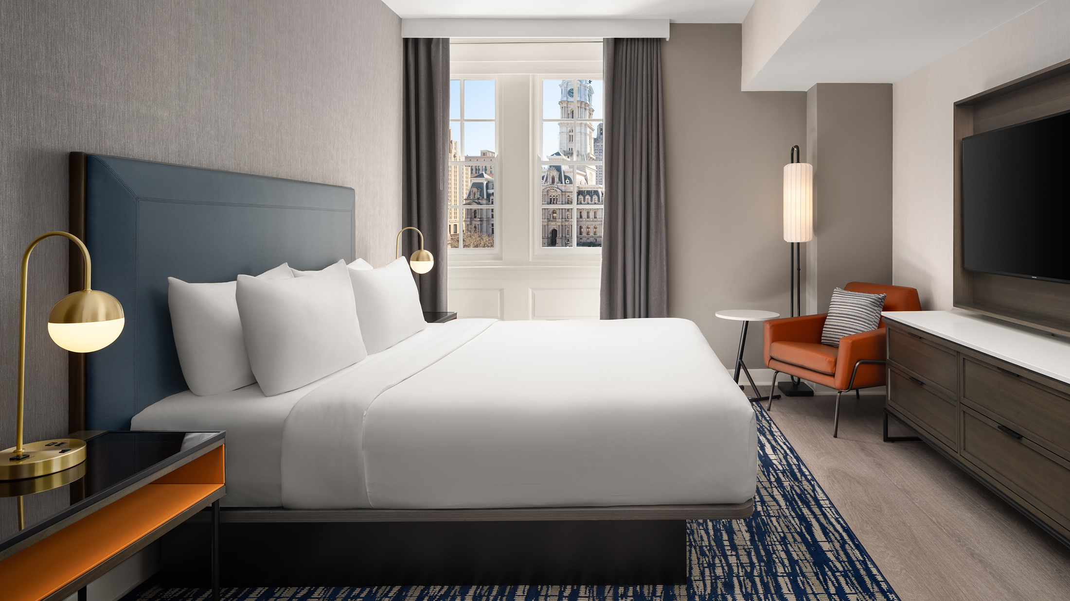 A hotel guestroom with a king bed with white linens, a orange accent chair and a window overlooking Philadelphia's City Hall.
