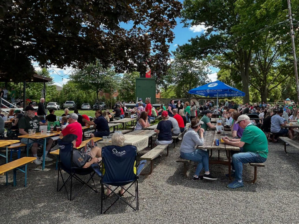 People sit at long picnic tables at Cannstatter Volkfest-Verein's biergarten, under string lights and green trees.