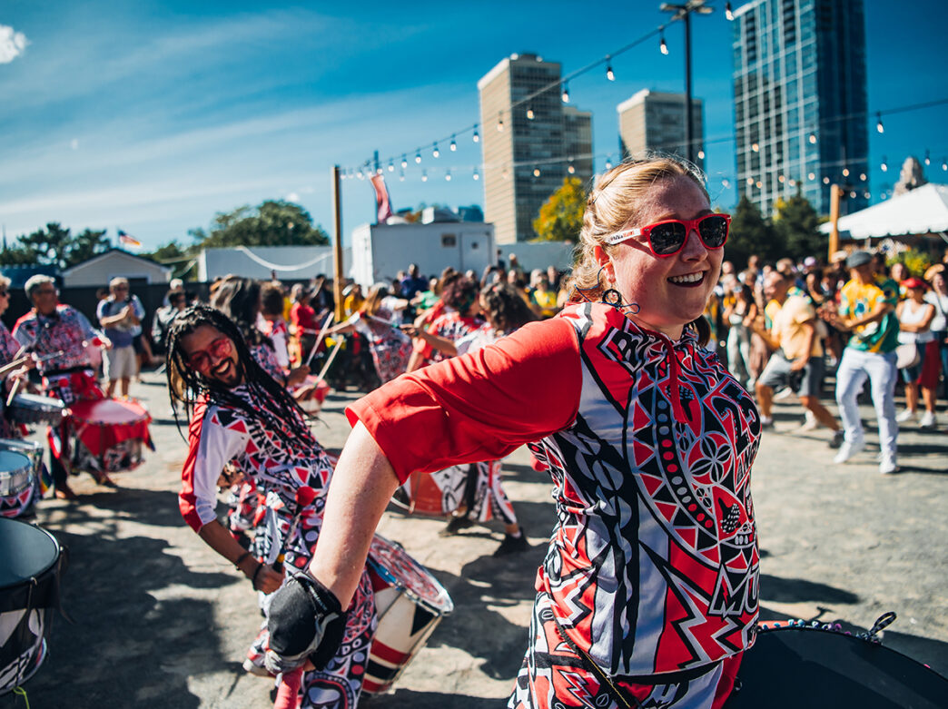Performers in red, black and gray outfits play the drums in front of a large crowd at an outdoor festival.