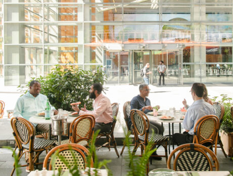 Two groups of people dine outdoors at Cafe Click in Philadelphia, enjoying meals at round tables surrounded by greenery and modern glass architecture.