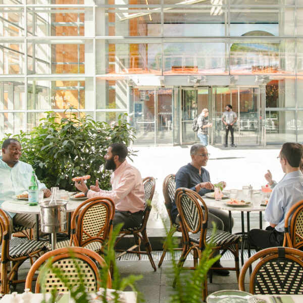 Two groups of people dine outdoors at Cafe Click in Philadelphia, enjoying meals at round tables surrounded by greenery and modern glass architecture.