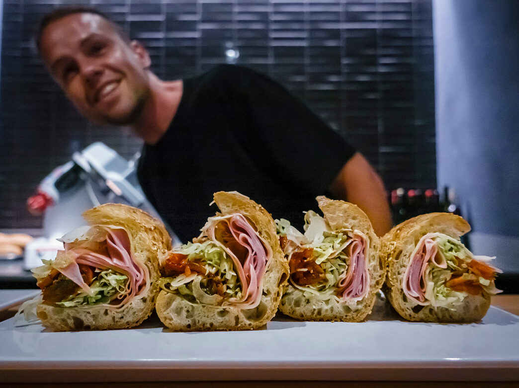 A chef smiles in the background as four neatly sliced hoagie pieces filled with ham, shredded lettuce, cheese and roasted peppers sit on a white platter at Pizzeria Beddia.