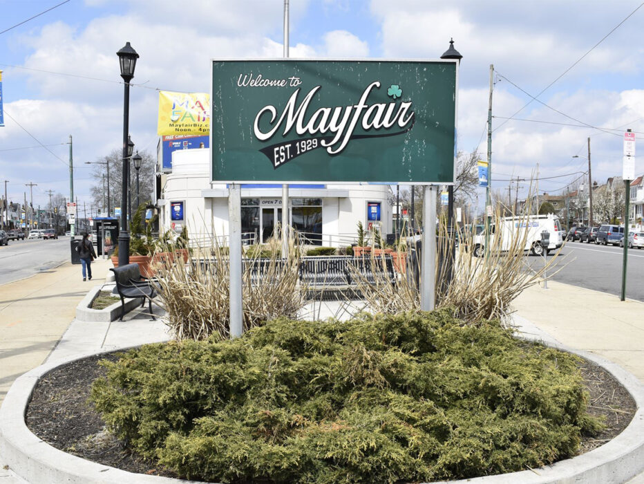 A green "Welcome to Mayfair" sign stands in a gardening bed surrounded by shrubbery at a intersection in Mayfair.