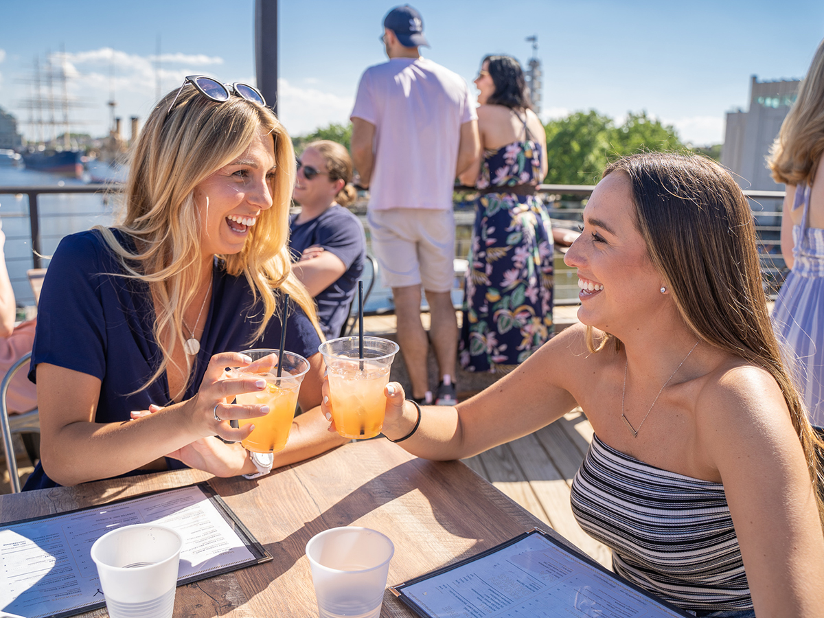 Two people smile and toast cocktails in clear plastic cups while dining outdoors at Liberty Point. Blue skies, menus on the table and the Delaware River are visible in the background.