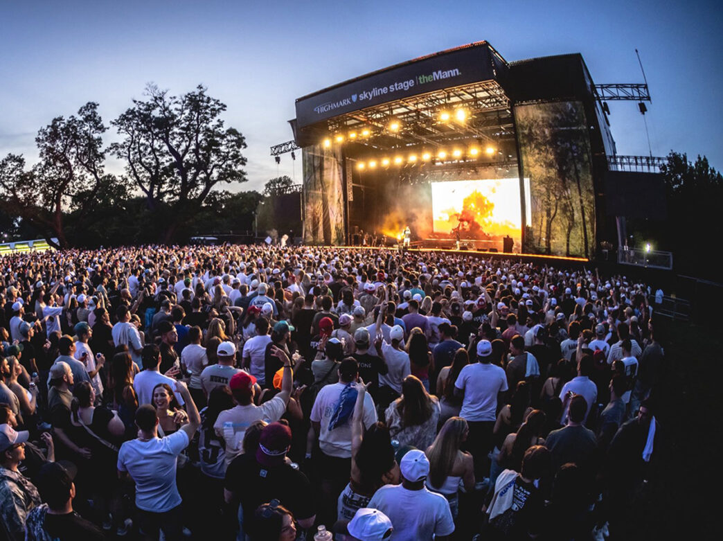 A large crowd gathers in front of a brightly lit outdoor stage at the Mann Center during an evening outdoor concert.