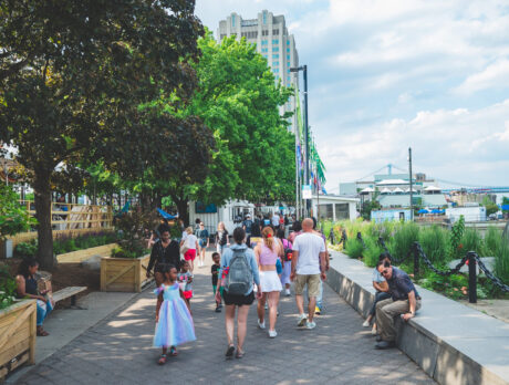 Visitors stroll along the waterfront path at Spruce Street Harbor Park on a sunny day. Trees line the path and the Benjamin Franklin Bridge is seen is the distance.