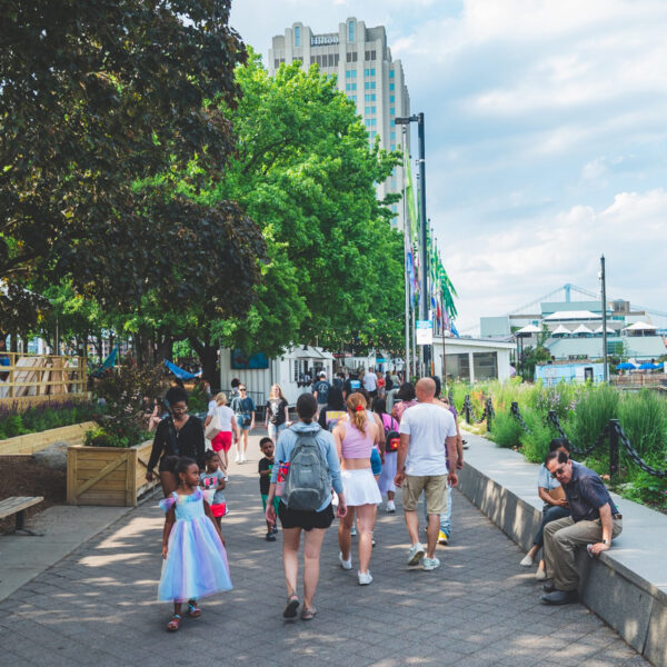 Visitantes passeiam pelo caminho à beira-mar no Spruce Street Harbor Park em um dia ensolarado. As árvores margeiam o caminho e o Benjamin Franklin Bridge é visto à distância.