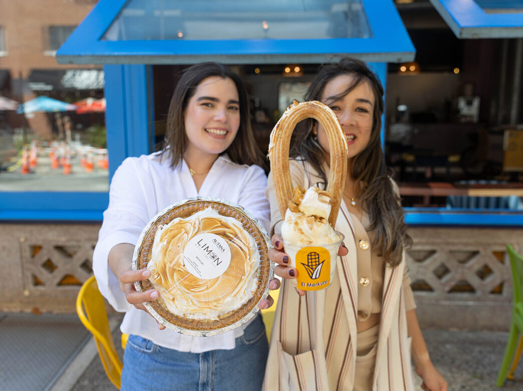 Two smiling women hold up desserts, one with a lemon meringue pie and the other one with ice cream topped with a churro.