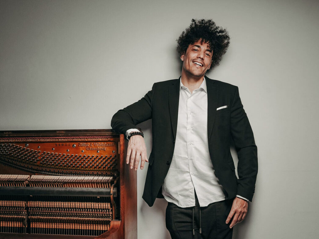 A man wearing a black suit jacket over a white button down shirt leans on an upright piano and smiles at the camera.