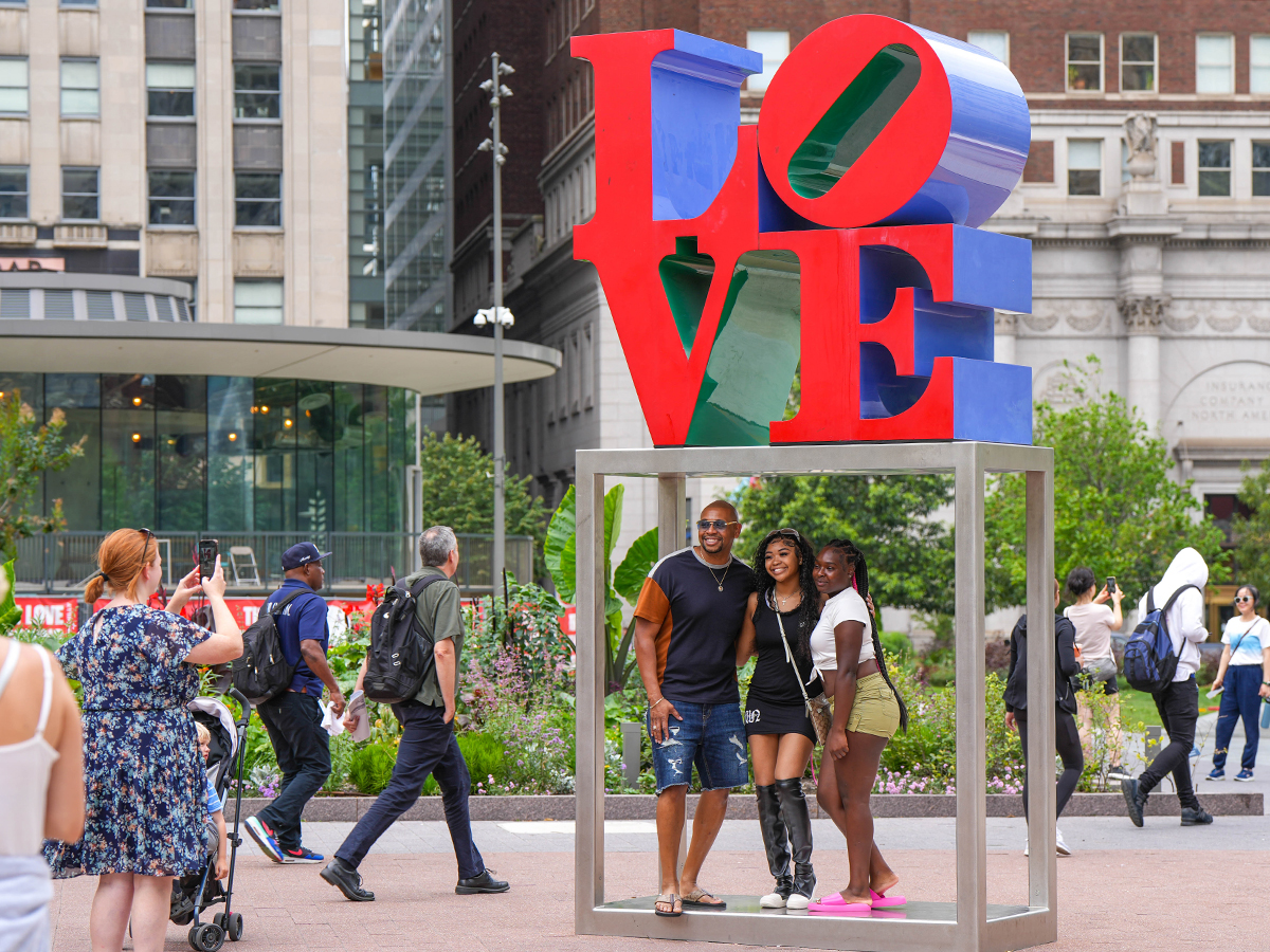 Visitors pose for photos under the iconic red and blue LOVE Statue, with people walking by and city buildings in the background.