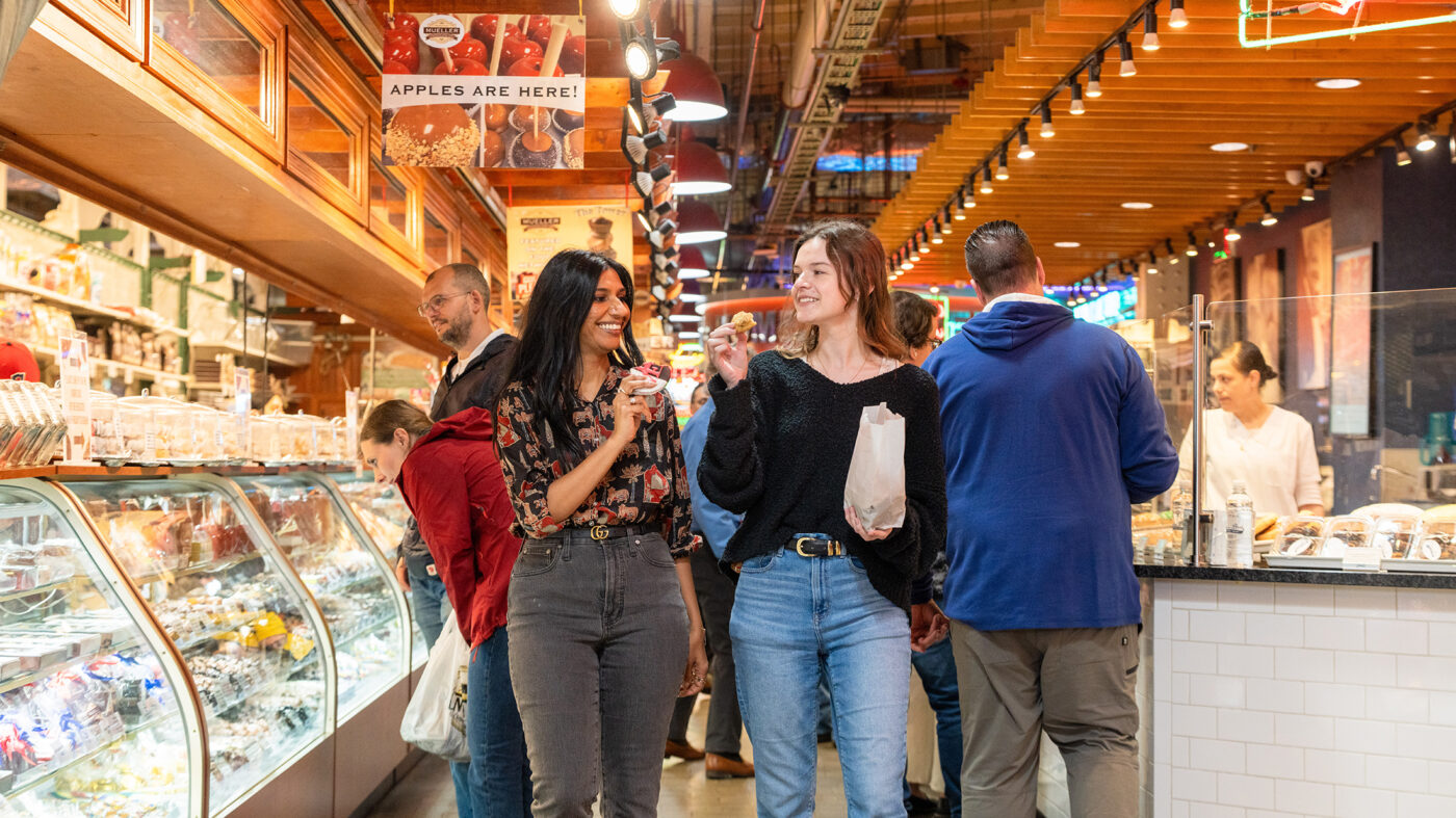 Two people smile while sampling treats inside the bustling Reading Terminal Market, surrounded by food counters and hanging signs.