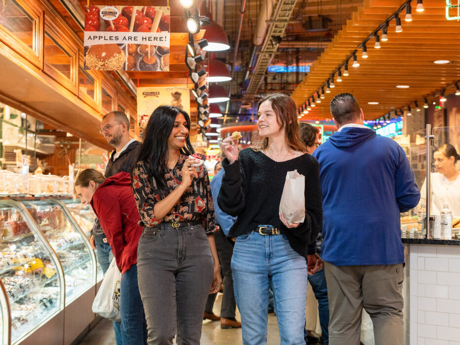 Dos personas sonríen mientras prueban delicias dentro del bullicioso Reading Terminal Market, rodeadas de mostradores de comida y letreros colgantes.